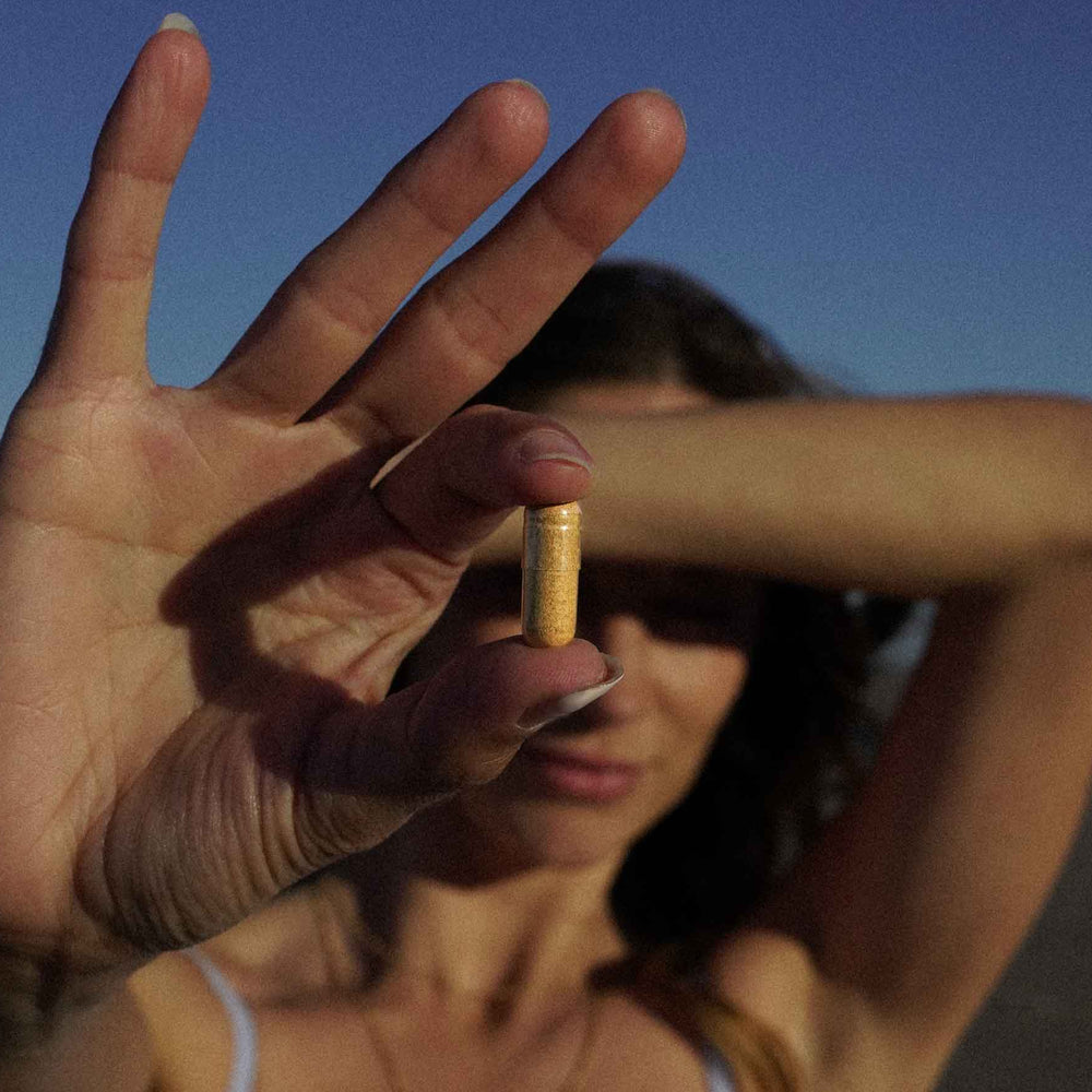 Woman on a beach holding a capsule in front of her face with a clear blue sky.