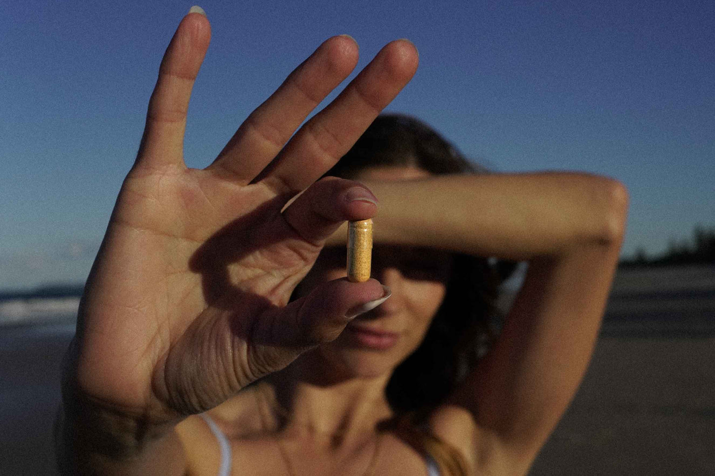 Woman on a beach holding a capsule in front of her face with a clear blue sky.